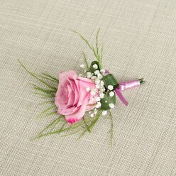 A romantic blush pink rose boutonniere is displayed against a neutral, woven beige fabric, reminiscent of natural linen you might see in the calm interiors of a Clerkenwell studio flat. The composition is photographed from slightly above, revealing how the rose sits in semi-open bloom, its velvety petals unfolding from a soft, almost porcelain pink at the center to a richer, more luminous pink at the outer edges, where they curl just enough to hint at movement and vitality. Encircling the rose, clusters of delicate white baby's breath create an ethereal, cloud-like frame, each tiny flowerhead adding lightness and a sense of gentle volume. Wisps of slender green foliage extend from behind the main bloom, their fine blades arching outward in relaxed, graceful lines, complemented by a single broader, dark green leaf that has been folded and wrapped to support the base like a bespoke collar around a shirtfront. The stems are carefully wrapped in a lustrous satin ribbon, blending shades of lavender-rose and deeper magenta-purple, the texture smooth and slightly reflective, as though catching soft afternoon light drifting in from a window overlooking Clerkenwell Road. The ribbon is tied in a neat, unfussy knot, conveying understated elegance rather than showiness. The background fabric's subtle weave appears under the boutonniere like a calm, textured canvas, making the pinks and greens stand out without distraction. This image evokes the quiet preparations before a wedding or prom, a small but meaningful floral accent ready to be pinned to a lapel for a ceremony in a nearby registry office or a stylish celebration in one of the area's converted warehouses, where modern design meets timeless romance.