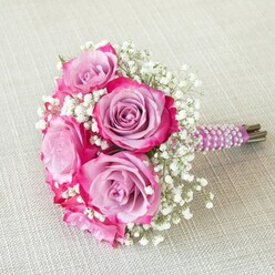 Echoing the unique vibrancy of Clerkenwell, this lavish bridesmaid bouquet features five radiant roses with dreamy lavender centres that dissolve into playful shades of fuchsia at the petal tips-a living palette that glows with movement and artistry. Crisp sprays of baby's breath-each tiny bloom like a dewdrop-are interwoven among the larger rose heads, imbuing a weightless, cloud-soft contrast that offers a sense of joyful celebration. The sophisticated handle is wrapped in deep purple satin, and detailed with winding strands of pearly white beads, a tactile flourish reminiscent of fine design found in Clerkenwell's creative studios along Rosebery Avenue. Resting gently on a lightly textured oatmeal linen, the bouquet's compact, rounded form is highlighted by soft daylight filtering in from a nearby window, creating soft shadows and dancing highlights across the arrangement. This bouquet's fragrance and colours together suggest fresh beginnings-its rich ribbon and pearl-wrapped handle the kind of thoughtful flourish a true local florist takes pride in. Perfect for couples marrying at Finsbury Town Hall or those planning a meaningful celebration nearby, this arrangement beautifully weaves together elegance, emotion, and the inimitable energy of Clerkenwell.
