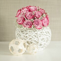 A serene floral composition captures a domed bouquet of pink roses and white baby's breath arranged in a sculptural white wicker-style sphere, in a setting that feels like a light-filled Clerkenwell loft near Exmouth Market. Around fifteen roses are gathered tightly at the top of the vase, their petals unfurling in layers of colour that move from a gentle, powdery blush at the heart of each flower to a more saturated, almost fuchsia pink along the outer edges. This natural gradient gives each rose a painterly quality, as if brushed with deeper colour at the tips. Between the roses, airy sprays of gypsophila introduce fine, white blossoms that soften the overall shape and create a delicate, halo-like texture. The bouquet is cradled by a round white vessel made from interwoven strands resembling painted rattan or wicker, its openwork design creating tiny windows where light slips through and dances onto the smooth, white tabletop beneath. Beside the vase rest two decorative spheres formed from pale, interlaced wooden strips, their neutral beige tones echoing the natural materials and adding a simple, Scandinavian-style touch. Behind everything is a softly textured wall in warm beige or cream, reminiscent of a carefully curated Clerkenwell interior, chosen to let the flowers' colours take centre stage without distraction. Soft, diffused lighting envelops the scene, revealing the velvety texture of the petals and the feathery structure of the baby's breath. The overall mood is one of modern romance and quiet refinement, ideal for marking special occasions such as anniversaries, Mother's Day, or a heartfelt thank-you delivered to homes and studios across EC1.