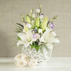 An elegant wedding-style centerpiece in white and soft lavender tones is captured against a calm, neutral background, much like a refined tablescape in a Clerkenwell loft venue. At the heart of the image stands a rounded white vase with an open lattice design, its woven, rattan-like texture creating a sense of lightness, as though the flowers are resting on air. From this sculptural base, tall green lily buds and fully open white lilies rise, their trumpet-shaped blooms wide and welcoming, each petal smooth and luminous in the soft, even light. Between these star-like flowers, lisianthus stems meander gracefully, some blooms showing pure white petals, others edged or brushed with gentle lavender-purple that deepens toward the centre. Several buds are still tightly wrapped, suggesting longevity and ongoing beauty. Small clusters of white filler flowers, similar to baby's breath, mingle with deeper purple or blue-toned accents, giving a delicate, almost whisper-like texture. Feathery fern fronds and fine, airy greenery spill slightly over the sides, balanced by broader, richer green leaves that anchor the arrangement visually. In the foreground, two decorative spheres made from pale woven strands lie on the pristine white tabletop, their open-weave structure echoing the vase and adding a modern, design-conscious touch. The backdrop is a light beige wall with a subtle, horizontal linen effect, softly lit to create a tranquil, timeless atmosphere. The composition suggests a bouquet perfect for wedding table arrangements near Exmouth Market or as a peaceful centrepiece for intimate gatherings in Clerkenwell, conveying quiet celebration, comfort, and refined taste.