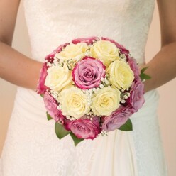 Bathed in gentle daylight reminiscent of Clerkenwell mornings, this evocative portrait spotlights a bride at her most radiant, her arms gently embracing a round bouquet meticulously assembled with closely nestled cream-yellow and vivid fuchsia roses. Each blossom is thoughtfully placed, playing with the tension between softness and vibrancy; several pink blooms reveal whispers of mauve, imparting a romantic depth reminiscent of local gardens in spring. Wisps of tiny white baby's breath are gracefully woven amidst the roses, adding feathery texture and subtle contrast that elevates the entire arrangement. Between the petals, slivers of deep green foliage ground the composition, inviting the senses to linger on the scent and freshness of expertly sourced Clerkenwell flowers. The bride's wedding dress, visible from the waist up, boasts delicate lace embroidery-a hallmark of vintage style that harmonizes with the elaborate floral work. The bouquet, luminous against the smooth, flowing gown, commands the viewer's focus, while the softly blurred, pale background hints at joyful celebrations nearby, be it at a Farringdon townhouse or the historic Charterhouse. This image exudes warmth and sophistication, offering an authentic glimpse into Clerkenwell's bridal artistry and the enduring significance of flowers in love stories celebrated across the local community.