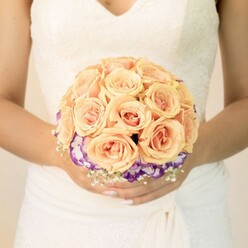 This image offers an intimate, waist-level view of a bride gently holding her bouquet, the kind of quietly emotional moment you might glimpse outside a registry office near Clerkenwell Green. Her gown is a traditional bridal white with a soft ivory cast, featuring a fitted lace bodice that tapers into a subtle V-neckline. The lace is intricate yet refined, echoing floral motifs that feel almost like they've been sketched in thread. Below, the skirt's fabric falls in smooth, graceful layers, adding a sense of movement even though she stands perfectly still. Centred in the frame is a hand-tied, round bridal bouquet, crafted with the care of an experienced florist who knows how to balance colour and texture. The upper portion of the bouquet is a generous cluster of roses in gentle, sun-warmed hues of peach, apricot, and blush pink. Each rose is tightly petalled, the outer edges soft and velvety while the centres deepen into warmer tones, creating an ombré effect that draws the eye inward. Around the base, a ring of ruffled purple blooms edged in white-likely lisianthus-curls outward, providing contrast and a rich, jewel-like accent. Tiny white baby's breath peeks between the larger flowers, adding lightness and a feeling of airy lace. The bride's hands, resting calmly around the stems, show a simple, pale manicure with a faint glimmer on one thumb, a small detail that mirrors the bouquet's understated glamour. The atmosphere is one of tender anticipation and classic romance, the kind of bouquet that would feel at home in wedding photographs taken along the cobbled streets and historic buildings that define Clerkenwell's character.