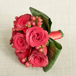A refined hand-tied bouquet of rich red roses and berries lies gently on a light beige, textured cloth, evoking the calm of a quiet Clerkenwell morning before the shops along Farringdon Road fully wake. The fabric beneath has a soft, woven grain that adds warmth without drawing attention from the flowers, allowing the saturated reds to glow. At the heart of the design, six or so roses unfold in layers of velvety petals, their deep crimson tones shifting to scarlet at the edges, like silk gently catching the light. Tucked between the blooms, small clusters of rounded Hypericum berries in shades of ruby and raspberry create a joyful rhythm, their smooth, slightly reflective skins offering a lively counterpoint to the roses' plush texture. Embracing the arrangement, broad, dark green leaves are artfully curled and folded, their strong lines and glossy finish adding structure and an almost architectural elegance, as if framing a miniature garden. A rich red satin ribbon winds tightly around the hidden stems, with a glimpse of its sheen visible at the side, hinting at the careful craftsmanship behind the bouquet. The lighting is soft and even, revealing the subtle shadows in the rose centres and the gentle highlights on the leaves without any glare, giving the arrangement a peaceful, timeless feeling. It is easy to imagine this piece being carried by a bridesmaid stepping out of a townhouse near Clerkenwell Green, or delivered by Clerkenwell Flowers as a romantic surprise, its fragrance mild but fresh, its colours speaking clearly of devotion, celebration, and heartfelt gratitude.