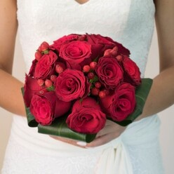 Capturing the romantic heart of Clerkenwell, this evocative photograph at once conveys the intensity and poetic grace of a traditional red rose bridal bouquet. Seen up close from the bride's perspective, the arrangement radiates drama-each velvety rose, from tight bud to lush blossom, arranged with masterful precision. The bouquet is strikingly compact, its rounded form held elegantly between hands with pale, freshly manicured nails. Between the vivid roses, clusters of rich ruby hypericum berries peek out, adding playful texture that echoes the vibrant hues seen in Clerkenwell's street flower markets or tucked away gardens near Spa Fields Park. A necklace of wide, dark green leaves serves as the structured base, their sheen polished as if touched by early sun filtering past Smithfield. The white wedding gown's intricate lace bodice and soft, flowing drape flutter in the periphery, providing a crisp backdrop for the burning reds of the floral composition. There is an almost tangible scent-sweet, almost heady-drawing out memories of secret springtime strolls along Rosebery Avenue. This bouquet does not just make a statement; it embodies unity, celebration, and enduring love, crafted for a bride who wants to blend classic romance with the urban edge of Clerkenwell. The richness of color, the careful balance of tradition and boldness, ensures this floral design will remain a vibrant, heartfelt part of any Clerkenwell wedding's cherished memories.