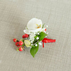 A detailed top-down photograph shows a single, handcrafted boutonniere laid on a softly textured, light beige linen or burlap-style fabric, reminiscent of the relaxed yet elegant interiors found in Clerkenwell loft venues. The focal point is a flawless white Phalaenopsis orchid, its rounded petals smooth and luminous, curving gently outward as if opening to the light. At its centre, a subtle wash of creamy yellow and soft orange glows from the orchid's throat, giving a gentle warmth that draws the eye inward. Surrounding the orchid, dozens of tiny white baby's breath blossoms cluster together, forming a delicate, cloud-like frame that introduces a sense of lightness and air, like mist drifting over nearby Spa Fields on an early morning. Tucked in close to the flowers, a cluster of glossy red berries adds a vibrant splash of colour, their polished surfaces catching the light and hinting at festive occasions or winter celebrations, while a few smaller green berry buds lend a note of freshness and natural variation. Interwoven among the blooms and berries, rich green leaves of slightly differing shapes-some broad and pointed, others more slender-give structure and balance to the arrangement, enhancing the contrast between the bright white blooms and the intense red fruits. The stems are neatly bound with a vivid red satin ribbon, its smooth, slightly reflective texture wrapped carefully and knotted with precision, creating a clean finish that suggests professional florist craftsmanship. This small but expressive floral piece feels ideal for a groom or guest at a Clerkenwell ceremony, a prom in central London, or a formal dinner around Farringdon, adding a touch of refined botanical style to a tailored lapel.
