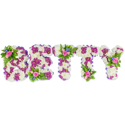 This bespoke sympathy tribute spells BETTY in strong, uppercase letters, each letter built from a tight, cloud-like mass of ivory-white chrysanthemums that give the whole piece a soft, tactile presence. Contrasting magenta-purple chrysanthemums are worked into the design to trace inner shapes and highlight edges, their rich hue lending a sense of depth and reverence. A discreet purple ribbon trims the sides of the letters, adding a neat, finished line that harmonises with the floral tones. Small, tender accent clusters punctuate the design-one tucked into the upper curve of the B, another situated on the top of the first T and a third on the upper arm of the Y-each cluster formed of soft pink roses, some in bud and some opening, accompanied by small sky-blue flowers (suggestive of delphiniums or cornflowers) and lush ferny greens that breathe life into the arrangement. The overall scent is gentle: the mellow perfume of roses balanced by clean, green undertones from the foliage and the powdery freshness of chrysanthemums. Photographed on a stark white background with even light, the colours read calm and respectful: white for peace, purple for dignity, pink for tenderness, and blue for remembrance. As a local florist might explain, this is a considered choice for services in Clerkenwell or for delivery to chapels near Exmouth Market-an elegant, heartfelt tribute that combines traditional sympathy symbolism with careful, contemporary floral craftsmanship.
