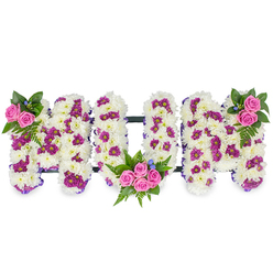 Soft daylight reveals a lovingly made floral tribute in the form of the word MUM, each capital letter sculpted from a thick mattress of immaculate white chrysanthemum blooms. The tiny, layered petals create a velvety, cloud-like surface that invites touch and quiet reflection. Throughout this white foundation, small groupings of magenta and deep purple spray chrysanthemums are thoughtfully placed to break the monotone with gentle rhythm and depth, their richer tones catching the eye without overwhelming the serenity of the piece. Three clusters of vivid pink spray roses act as visual hearts: a modest pair at the top left of the first M, a larger, more generous cluster nesting in the lower curve of the U, and a mirrored cluster atop the final M. These roses, with their fresh, slightly fruity scent and tightly cupped petals, are framed by lush green fern leaves and mixed foliage that lend graceful movement and architectural balance. Tiny blue accent blooms are dotted among the leaves, offering a cool counterpoint to the warm pinks. A slim dark bar connects the letters, while a delicate purple ribbon traces the outer edges, accentuating the shape. The overall mood is one of tender dignity - a handcrafted sympathy piece meant to comfort mourners and honour a mother's memory. A Clerkenwell floral studio would recommend this design for local services around Farringdon Road and St John's Square, where a thoughtful tribute like this helps families convey profound affection and respect.