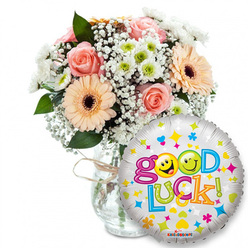 This image reads like a floral hug from Clerkenwell: a full, pastel-hued bouquet arranged in a clear glass vase with a rough twine bow, placed slightly left of centre on a white background. The flowers are chosen for comfort and optimism - soft pink roses with concentric, velvety petals, peach-orange Gerbera daisies whose darker centres give contrast and a sunny, tactile quality, airy clouds of white baby's breath woven through like bridal lace, and tight sprays of white and lime-green chrysanthemum florets that lend a crisp, lively note. Glossy dark-green foliage peeks through, framing the blooms and adding relief to the pale palette; imagine the satiny feel of rose petals against the firmer daisy heads, and the light citrusy lift the chrysanthemums bring. Crowding playfully in the foreground to the right is a round silver foil balloon, its metallic sheen catching a soft studio light and reflecting the bouquet's colours back in miniature. The balloon is decorated with scattered hearts, stars and tiny colourful shapes, and proclaims "Good Luck!" in a bouncy, multicoloured font - the two 'o's in "Good" replaced by vivid yellow smiley faces, one winking, one sticking out its tongue - which brings a cheeky, human touch to the sentiment. A small Kaleidoscope logo sits near the balloon's base. This composition feels handcrafted and kind - a perfect Clerkenwell pick-me-up for new starts, exams or farewells, easily imagined being prepared and locally delivered by a neighbourhood floral maker around St. John's Gate or Farringdon Road.