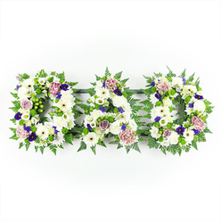 A horizontal floral tribute spelling out DAD in three distinct block letters is displayed against a clean, bright white background, allowing every nuance of the design to stand out clearly. Each letter is carefully built from a dense bed of white chrysanthemums and white gerbera daisies, their rounded petals overlapping to form a soft, cloud-like surface that speaks of purity, remembrance, and quiet love. Interwoven among these are elegant white lisianthus and small white spray roses, giving the arrangement a slightly ruffled, garden-fresh texture. Lush green foliage, dominated by graceful fern fronds and sprigs of glossy greenery, outlines and cradles each letter, providing a rich contrast that strengthens the definition of the word. Here and there, clusters of bright green button chrysanthemums and hypericum-style berries add tiny points of interest that draw the eye along the curves of the D and the crossbar of the A. Deeper colour notes come from violet and royal purple flowers, similar to petite pansies or violas, along with mauve roses and dusky pink carnations that seem to glow gently in the soft, even light. The tribute feels as though it has just been arranged in a calm studio near St John Street in Clerkenwell, ready to be delivered to a local church or crematorium. Its mood is respectful yet deeply personal, a bespoke sympathy arrangement that lets family members express their love and gratitude for a father's life during a funeral, memorial service, or private farewell.