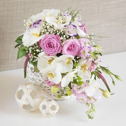 An artfully styled floral centerpiece is captured in soft, natural light, as if set on a wedding table in a converted Clerkenwell warehouse, its details revealed in a close, intimate view. A low, round basket made from white-painted wicker forms the base, its tight weave and subtle texture giving a relaxed yet polished character. From this basket, a generous arrangement of fresh flowers spills outward in shades of rose pink, creamy white, and gentle lilac. Fully-opened pink roses sit prominently at the centre, each bloom rich with layered petals that curl into perfect spirals, evoking romance and quiet affection. Elegant white Phalaenopsis orchids arc gracefully through the design, their smooth, waxy petals and golden-yellow centres adding an exotic, modern note. Around them, ruffled white Lisianthus edged with hints of lavender-purple introduce softness and depth, while pale pink stock stems extend slightly beyond the main body, creating movement as they cascade over the rim. Fine sprays of baby's breath are scattered throughout, like a mist of tiny white stars, giving the bouquet a light, airy texture that contrasts beautifully with the denser roses and orchids. Fresh green foliage is carefully tucked between blooms, with some long leaves and unopened buds slipping naturally to the sides, suggesting growth and life. The basket is set on a pristine white surface, beside two decorative balls woven from pale natural fibres, their interlaced strands echoing the basket's weave and adding sculptural interest. Behind, a softly textured beige or taupe fabric backdrop-similar to linen you might see in a design studio off St John Street-keeps the focus on the flowers. The arrangement conveys a sense of celebration, ideal for wedding receptions, anniversary dinners, or refined gatherings across Clerkenwell.