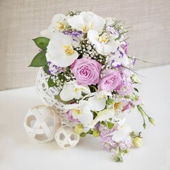 A refined floral centerpiece in soft whites, pinks, and purples is presented on a clean white tabletop, the kind you might find laid for a wedding breakfast in a converted Clerkenwell warehouse near St John Street. The flowers rise from a distinctive white spherical vase, its open, woven pattern reminiscent of fine wicker artfully shaped into a perfect globe. At the forefront, several white Phalaenopsis orchids open in serene arcs, their broad petals smooth as porcelain and their yellow-gold centres adding a hint of warmth and luxury. Nestled among them, pastel pink and lilac roses are captured at full bloom, with ruffled petals layered tightly at the heart and softening towards the edges, conveying a sense of quiet romance. Between the roses and orchids, smaller ruffled blossoms, likely Lisianthus, show white petals tinged with cool purple, bringing a subtle painterly quality to the design. Delicate sprays of white baby's breath weave through the bouquet like a fine mist, creating a light, airy texture that softens the silhouettes of the larger flowers. Slender stems of pale purple stock or similar trailing blooms extend down the side of the vase, forming a gentle cascade that draws the eye downward in a graceful curve. Fresh green foliage is tucked here and there for contrast, framing the pastel tones and adding depth. On the white surface beside the vase, two creamy woven decorative balls, one medium and one small, echo the basket-like pattern of the container and complete the composition. A neutral beige background, softly lit and slightly blurred, allows every petal, stem, and woven detail to stand out with clarity, evoking a mood of thoughtful celebration and modern elegance typical of a carefully styled Clerkenwell event.
