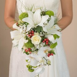 Cradled in a bride's hands just steps from Clerkenwell Green, a breathtaking bridal bouquet reveals a story of local artistry and romance. At its core, shining under gentle natural light, rests a swirl of creamy-white rose petals-emblematic of enduring love. Radiating outwards, grand white oriental lilies open wide, each petal adorned with faint rust-toned speckles, their rich fragrance suggestive of an early spring morning at Smithfield Market. The bouquet comes alive with vivid contrasts: vivid lime-green pom mums and cushiony hydrangea accents nestle among the blooms, their brightness offset by clusters of glossy red hypericum berries that lend a joyful, celebratory flair. Fluffy sprays of baby's breath float around the composition, their wispy presence softening the transition from bold colour to airy lightness. Trailing elegantly, phalaenopsis orchids extend in a graceful cascade, their pristine blooms capturing movement and sophistication, echoing the classic elegance often sought by couples marrying near Clerkenwell's historic churches. Fine stems of foliage, including slender, grass-like sprigs, provide vibrant structure, framing the arrangement with a subtle touch of the wild. Soft lace detail on the bride's gown and the gentle curve of her hands add an intimate charm, while the background blurs into subtle pastel tones, focusing every eye on the flowers' splendour. This Clerkenwell-inspired bouquet, radiant and fresh, offers a blend of modern style and traditional grace-perfectly reflecting the neighbourhood's mix of heritage and contemporary flair, and ideal for a memorable wedding day.