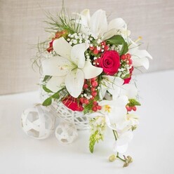 A sophisticated floral centrepiece in pure white and deep crimson rests on a spotless white surface, bathed in gentle, even light that picks up every curve and petal. The arrangement is cradled in a round, white container crafted with an airy, open weave, reminiscent of modern wicker or handworked rattan, giving the design a light, contemporary feel that would sit beautifully on a wedding table in Clerkenwell's warehouse-style venues. From this basket, several magnificent white oriental lilies rise and unfurl, their broad petals and exposed stamens creating a sense of calm drama, like a still moment before the toasts begin. Nestled between them are sumptuous red roses, their velvety petals tightly layered, suggesting timeless romance and quiet passion. Sprigs of delicate white baby's breath float between the larger blooms, adding a gauzy softness, while fine green foliage introduces a clean, fresh contrast that feels as crisp as an early morning walk around St John's Square. Glossy red hypericum berries are dotted throughout the design, glinting like tiny jewels and echoing the roses' rich tone. To the right, a cascade of white phalaenopsis orchids spills elegantly from the main bouquet, each bloom curved and poised, with subtle touches of yellow and blush at the heart that add depth without breaking the serene palette. On the left side of the image, a trio of white woven spheres, each slightly different in size, sits on the table, mirroring the basket's texture and tying the composition together. This arrangement captures the mood of a refined Clerkenwell wedding or anniversary dinner, combining modern design with classic romance in a way that feels both curated and effortlessly natural.