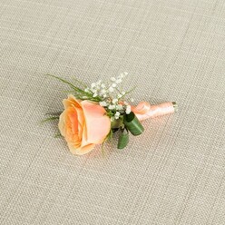 The image captures a delicately crafted peach rose boutonniere lying horizontally across a neutral, oat-coloured linen fabric, the fine weave of the cloth adding a calm, tactile backdrop. At the heart of the arrangement sits a single rose, partially open and carefully angled, its petals deepening to a warm apricot at the centre and softening to creamy peach along the outer edges. This gentle gradient makes the bloom appear luminous, like sunrise over the rooftops of Clerkenwell. Nestled just above and around the rose are clusters of white baby's breath, their tiny, cloud-like flowers forming a soft halo that emphasises the rose's fuller, velvety form. Wisps of slender green foliage, along with a couple of broader, darker green leaves, emerge from beneath the bloom, curving gracefully to give the buttonhole structure and a natural, garden-fresh feel. The stems are tightly bound with a matching peach satin ribbon, its smooth, matte surface wrapped in precise coils and finished with a discreet knot that keeps the profile sleek enough for a tailored suit lapel. The light is diffuse and even, revealing the delicate textures of each petal, the fine sprays of gypsophila, and the understated sheen of the ribbon without distracting glare. The overall mood is serene and romantic, evoking the quiet moments before a wedding at a local venue near Exmouth Market or a formal dinner along St John Street, where such a boutonniere would be pinned in place to mark a day of promise and celebration.