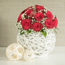 A striking, contemporary floral centrepiece is captured as though it were already placed on a wedding table in Clerkenwell, ready for guests to arrive. The focal point is a domed cluster of rich crimson red roses, each flower at the peak of its beauty, with layered petals curling inwards like soft satin ribbons. These roses form a tight sphere of colour, suggesting passion and commitment, the kind of arrangement often chosen for romantic receptions and civil ceremonies. Between the roses, clouds of white gypsophila sprinkle lightness throughout the design, their tiny blossoms catching the eye like flecks of lace against the bold red. Small touches of green foliage deepen the sense of freshness, as if the bouquet has just been prepared by hand in a nearby studio. The arrangement is cradled by a distinctive white vase with a rounded, bulbous shape and an open, interwoven structure reminiscent of painted wicker or sculpted branches. The negative spaces within the weave allow soft light to filter through, adding texture and gentle shadow to the composition. On the left of the vase sit two decorative cream-toned spheres made from similar woven strips, one noticeably larger, echoing the tactile, crafted feel that Clerkenwell is known for in its design shops and studios. They rest on a pristine white tabletop that reflects a touch of light, while a neutral, slightly textured wall forms a calm backdrop, blurred just enough to keep the roses in sharp focus. The scene suggests a quiet moment before a celebration, perfect for illustrating romantic table flowers or same-day wedding delivery across Clerkenwell and the surrounding streets.