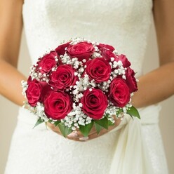 In the heart of Clerkenwell, where history and modernity caress each corner, a bride stands poised and tranquil, her hands wrapped lovingly around a lush bridal bouquet crafted with local expertise. The bouquet's centrepiece is a vibrant gathering of deep red roses, each bloom expertly shaped, velvet petals swirling inward to capture the rich intensity of classic romance. Interlaced among the roses, delicate clusters of pure white baby's breath flicker like gentle stars, their tiny petals forming a soft, airy mist that tempers the boldness of the crimson blossoms. Emerald leaves peek from beneath, framing the bouquet with the same crisp vitality seen in springtime strolls near St John's Gate. The bride's fingers, elegantly manicured, form a tender embrace around the stems, embodying the reassurance and anticipation of the moment. Her elegantly textured gown, perhaps woven with intricate lace reminiscent of Clerkenwell's centuries-old textile traditions, shimmers subtly beneath the bouquet and is complemented by a delicate sash trailing gracefully to the side. The background is a wash of warm, neutral light, drawing the eye endlessly back to the bouquet and the bride's poised expression. Imbued with the artistry and freshness that defines Clerkenwell Flowers, this composition becomes more than just a floral display-it narrates a story of enduring love, style, and gentle anticipation echoing through every bloom and petal.