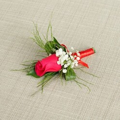 A carefully crafted boutonniere is photographed against a neutral, light beige textile background, the fabric's gentle texture suggesting linen or fine burlap, like something you might find in a stylish Clerkenwell townhouse near St John Street. Centered in the composition is a single rosebud, its petals a vivid, luscious shade of pink that leans toward fuchsia, with deep magenta tones hidden in the tight spiral of the bloom. The rose is still mostly closed, promising full beauty yet to unfurl, which lends a sense of new beginnings that feels perfect for a wedding morning or a milestone celebration. Surrounding the rosebud are soft tufts of white baby's breath, each tiny flower forming part of a delicate collar, creating a cloud-like contrast that enhances the rose's saturated colour. Wispy sprays of bright green, grass-like foliage extend outward, their fine, feathery lines adding lightness and movement, while slightly broader green leaves tuck in at the base, giving the piece balance and structure. The stems are gathered tightly and wrapped in a smooth coral-pink satin ribbon, its sheen gently reflecting the light in subtle, festive glimmers. The ribbon sits snug and precise, with no stray ends, suggesting the steady hands of an experienced florist used to preparing buttonholes for ceremonies across Clerkenwell and nearby Farringdon. The bright, even lighting reveals the velvety texture of the rose and the freshness of the greens, while the neutral background keeps all attention on the delicate interplay of colour and form, evoking a sense of poised elegance and thoughtful craftsmanship ready to be worn on a lapel.