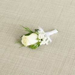 This image presents a classic wedding boutonnière laid on a diagonal across a pale, linen-like fabric, whose subtle diagonal weave and warm beige tone create a calm, neutral backdrop. At the centre is a single ivory rosebud, its petals plump, velvety, and tightly layered, with the outermost petals just beginning to curl away from the core. The creamy white tone of the rose is soft rather than stark, picking up the warm light that gently grazes the arrangement. Tucked around the rose, tiny clusters of white baby's breath form a halo of miniature blossoms, like floating flecks of cloud that build a sense of volume without overwhelming the main flower. A few slender, dark green leaves stretch from beneath the bud, their surfaces smooth and slightly glossy, adding structure and contrast. Interspersed are finer fern-like fronds that introduce a delicate, feathery element, reminiscent of greenery gathered from a shaded garden courtyard off Clerkenwell Green. All of the stems are gathered and expertly wrapped in a crisp white satin ribbon, wound tightly and evenly to form a sleek handle, then finished with a small, neat bow positioned just beneath the rose. The ribbon's soft sheen catches the light, hinting at the touch of a skilled florist preparing pieces for a ceremony in nearby venues along St John Street or around Farringdon. The lighting is gentle and diffused, with no harsh highlights, emphasising texture-the weave of the fabric, the satin of the ribbon, the velour of the petals. The composition, with its restrained palette of whites and greens, conveys understated refinement, ideal for grooms, groomsmen, or formal occasion wear in central London's Clerkenwell district.