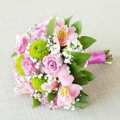 This bouquet, artfully composed on a creamy woven surface suggestive of Clerkenwell's eclectic studios, captures the poetry of spring in full bloom. At its heart rest soft, blush-pink roses, their layers opening in delicate swirls, each blossom a symbol of timeless romance and local heritage. Bright green button chrysanthemums add a burst of lively colour and tactile intrigue, echoing the playful vibrancy found in Clerkenwell's flower stalls on Leather Lane. Pink alstroemeria weave gracefully through the arrangement, their slender petals adorned with sunlit gold streaks and speckled accents-each a brushstroke in a living canvas. Airy clusters of baby's breath give the bouquet a light, ethereal quality, reminiscent of gentle Clerkenwell mornings when the air is fresh and full of hope. Verdant leaves and filler greenery wind through the design, grounding the composition and enhancing its organic character. Everything is drawn together with a rich magenta ribbon, whose satin sheen subtly complements the floral palette and adds a final note of sophistication. The bouquet emanates a fresh, bright energy under soft light, conjuring memories of new beginnings and heartfelt celebrations. Whether gifted for a wedding at St James's Church or a special anniversary near Farringdon, it is a bouquet that breathes with love, memory, and the promise of another beautiful season in Clerkenwell.
