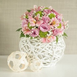 On a pristine white surface, a lush floral centerpiece rises like a soft pastel cloud, set against a gently textured beige backdrop that recalls the warm, muted walls of an elegant Clerkenwell apartment near Spa Fields. The bouquet is densely packed yet carefully composed, overflowing with pastel pink and lavender roses at their fullest, their velvety petals unfolding in generous spirals. Nestled among them are pink alstroemeria, each slender bloom marked with fine stripes and flecks of darker pink and soft yellow, adding a subtle sense of movement and detail when you imagine tracing the petals with your fingertips. Bright lime green button chrysanthemums dot the arrangement with vivid, round pops of colour, introducing a crisp, modern contrast that keeps the design lively rather than overly sweet. Fine clouds of delicate white baby's breath drift through the flowers, filling small gaps and lending an airy, romantic haze to the whole display, while rich dark green leaves offer an anchoring backdrop of natural depth. All of these elements are gathered into a distinctive white vase crafted with an open, woven lattice of narrow strips, its rounded, weighty shape suggesting sturdiness beneath the intricate surface. To the left, on the same white tabletop, rest two decorative woven spheres in pale, natural tones, their open rattan-like structure mirroring the vase and creating a harmonious trio of textures. The soft, even lighting highlights every curve and petal, evoking an atmosphere of quiet joy and gentle celebration-just the sort of centerpiece you might choose from a local Clerkenwell florist to brighten a birthday brunch on St John's Square or a relaxed anniversary dinner at home.