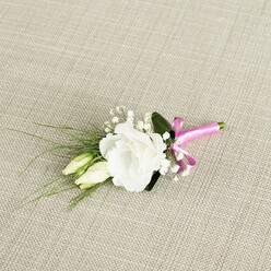 A refined floral boutonnière lies diagonally across a warm, oatmeal-coloured fabric backdrop, the gentle weave echoing the tactile feel of natural textiles often seen in Clerkenwell's artisan studios. The central focus is a single white eustoma, or lisianthus, fully open and perfectly formed, its softly ruffled petals layered in concentric waves that create a tender, luminous heart. Just to the left and slightly below, two elongated buds of the same flower variety rest close to the main bloom, their pale green and cream tones suggesting the quiet promise of blossoms still to come, like a wedding morning building towards the vows. Threaded between them are fine stems of white gypsophila, their tiny, cloud-like flowers introducing a delicate, airy fullness that softens the overall outline. From behind the central lisianthus, slender blades of deep green, grass-like foliage arc out towards the lower left, adding movement and a subtle sense of wildness, balanced by a single broader, darker green leaf that anchors the design. At the base, the stems are firmly bound with a vivid fuchsia pink satin ribbon, tied into a compact bow whose silky texture catches the light. This bold ribbon provides a joyful accent of colour against the muted neutrals, suggesting personality and celebration without overpowering the gentle palette of whites and greens. Photographed under soft, even lighting, every curve of petal and touch of greenery is visible, evoking a calm, romantic mood-ideal for a groom or guest heading to a ceremony near St John Street or one of Clerkenwell's historic townhouses, carrying this small piece of floral artistry close to the heart.