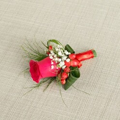 A single, impeccably arranged boutonniere rests against a pale beige, linen-like cloth, the kind of neutral, textured fabric often used on tables in stylish Clerkenwell venues. At the centre of this small floral piece is a striking fuchsia rose, its petals dense and velvety, still in that perfect stage where the bloom is open yet full of life, with the inner whorls curling protectively around its heart. Around the base of the rose, delicate sprays of white baby's breath gather like tiny stars, their airy flowers bringing a soft, clouded halo that contrasts beautifully with the strong pink tones. Tucked between the white blooms are clusters of bright red hypericum berries, each one smooth and glossy, providing a joyful pop of colour that feels at once festive and modern. Embracing the flowers is a mix of greenery: broad, dark green leaves sculpted and slightly curled to form a neat, tailored outline, and fine, fern-like foliage that spills gently outward, lending movement, texture, and a touch of wild garden charm. The stems are carefully wrapped in a strip of red satin ribbon, wound tight and straight so it lies flat against the fingers, the ribbon's sheen echoing the richness of the berries and making the whole design feel luxurious yet comfortable to wear. Gentle, diffused daylight glides across the arrangement, enhancing the vivid colours without harsh shadows, so each detail reads clearly. The boutonniere feels ideal for a groom, groomsman, or prom guest wandering through the cobbled streets near Charterhouse Square, carrying a piece that quietly speaks of romance, celebration, and the meticulous craft of a local florist who understands urban elegance and heartfelt occasions.