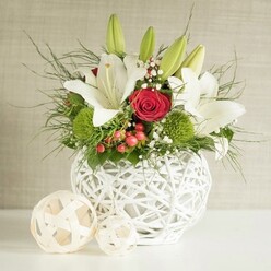 On a smooth white tabletop, a refined floral centrepiece sits in a striking round vase that looks as if it has been woven from slender white branches, its open latticework lending an airy, modern elegance reminiscent of contemporary interiors around Clerkenwell. The vase brims with lush, fresh flowers: tall stems of white lilies rise gracefully, some blossoms fully open with wide, star-shaped petals and warm yellow stamens, others held in tapered green buds that hint at fragrance still to unfold during an evening reception. Nestled amongst the lilies are rich red roses, their velvety petals spiralling inward to form deep, romantic hearts, evoking the intimate charm of a wedding breakfast in one of the converted warehouses off Clerkenwell Road. Bright green pom-pom blooms, likely dianthus or chrysanthemums, offer a soft, tufted texture and a vivid colour contrast, creating a lively dialogue between classic romance and contemporary design. Delicate baby's breath threads through the bouquet, its tiny white flowers forming a gentle mist around the bolder blooms, while clusters of red berries peek out, adding a touch of festive cheer suitable for autumn and winter celebrations. Various greens, from fine, feathery fronds to broader, glossy leaves, spill outward in relaxed arcs, giving the arrangement a natural, slightly untamed edge that balances its polished look. To the left of the main vase, two decorative spheres made from pale, woven material mirror the vase's pattern and provide a calm visual echo. The neutral, softly textured background keeps the focus firmly on the crisp whites, rich reds, and fresh greens, creating a centrepiece that feels timeless and perfectly suited to wedding tables and special occasions across Clerkenwell.