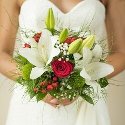 In the heart of Clerkenwell, this bridal bouquet unfolds like a love letter in florals, styled for a celebration in one of London's most creative quarters. The camera lingers on the bride's elegant, textured white dress-its modern simplicity providing a graceful backdrop. Cradled in her hands, the bouquet is alive with meaning: white lilies, some stretching open to reveal golden hearts, others tucked in as emerald-green buds, symbolize purity and fresh beginnings. Crimson roses, their velvety spirals tightly swirled, are nestled intimately among the lilies, their deep hue adding timeless romance to the day. A gentle scatter of baby's breath brings a sense of dreamy weightlessness, floating across the surface like memories caught in the morning breeze of Spa Fields. Vivid red hypericum berries peek out with festive charm, while sculptural green button blooms lend a playful, tactile surprise. Mixed greenery-from feathery fern tips to glossy leaves-anchors the palette and echoes the verdant parks and gardens surrounding Clerkenwell. The result is a hand-tied arrangement as dynamic as the area itself, full of textural contrast and delicate scent. Each element is chosen with care, crafting an arrangement that feels both classic and alive with contemporary artistry, tailored for a wedding morning that will be remembered for years along the streets and squares of Clerkenwell.