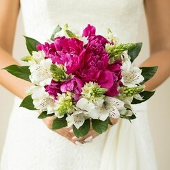 In this detailed bridal close-up, a vibrant hand-tied bouquet is held just above the waist of a bride, her white dress forming a gentle, textured backdrop like the pale stone walls around Clerkenwell Green. The bouquet is strikingly round and full, dominated by generous magenta and fuchsia peonies whose petals overlap in soft, ruffled waves, almost like layers of silk tulle. Nestled between the peonies are clusters of white alstroemeria, each blossom with elegant, elongated petals and finely speckled maroon markings running deep into the throat of the flower, adding a refined, almost painterly detail. Sharp, upright green buds and spiky filler stems pierce through the rounded form, providing lively contrast and giving the arrangement a sense of movement and freshness, as though it has just been gathered from a city garden after rain. Around the outer edge, broad, deep green leaves with a natural sheen cradle the flowers, framing the colour and lending structure to the bouquet. The bride's hands, relaxed and steady, cup the stems carefully wrapped in ribbon, her nails finished with a soft, understated pink polish that hints at quiet elegance. The lighting is bright yet gentle, bringing out the vivid interplay of magenta, white, and green without harsh shadows. The background remains softly blurred, ensuring every nuance of the flowers and fabric stands out. It feels like an intimate moment captured just outside a Clerkenwell registry office or converted warehouse venue, a bouquet crafted with care and artistry, ideal for inspiring couples planning a colourful London wedding in the heart of EC1.