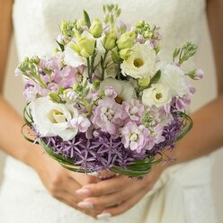 This bridal bouquet, inspired by the tranquil charm of Clerkenwell, radiates contemporary romance in every petal. Captured in a close-up, the arrangement appears freshly gathered-creamy lisianthus flowers, soft pink and lavender stock, and hints of unopened green buds come together in pastel harmony. The vivid purple allium at the base forms a striking halo, their spiky forms contrasting gently with the rounded, lush blooms above. Wisps of deep green bear grass spiral artfully, encasing the bouquet and emphasizing the careful craftsmanship typical of Clerkenwell's finest florists. Delicately manicured hands cradle the bouquet's stem, pale polish glowing in soft natural light, while the suggestion of a white lace wedding gown lingers in the blurred background-perhaps on a sunlit morning near Charterhouse Square. The overall composition is a testament to both the emotional vibrancy of wedding celebrations and the understated elegance central to modern Clerkenwell style: a bouquet that brings together fragrance, tenderness, and quiet sophistication, perfect for a spring or summer ceremony in the heart of London.