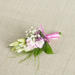 Set against a finely woven, light beige fabric that recalls the soft textiles of Clerkenwell design studios, this floral buttonhole is presented as though waiting on a suit jacket laid out for a city wedding. The corsage-style piece is a compact yet intricate arrangement, no more than a small handful of stems, but crafted with the care of an experienced local florist. At its heart sit several soft pink, multi-petalled blooms, their ruffled petals layered so densely they form a cushion of colour, gently shifting from pale blush at the centre to a slightly deeper pink at the edges. Nestled among them, tiny white baby's breath flowers float like a delicate mist, their many small florets adding airiness and a sense of movement. Dotted here and there, miniature sprigs of lavender or deep purple blossoms introduce a quiet contrast, the cooler tones deepening the overall palette without overpowering the gentle pinks and whites. To one side, a line of elegant, elongated buds in creamy white and fresh pale green, likely freesia or similar slender blooms, lie closed, suggesting fragrance and future bloom, as if they will open slowly during a reception on St John Street. Beneath the flowers, broad, glossy green leaves fan out to provide a lush, structured base, while wispy strands of fine, grass-like greenery extend beyond the main cluster, softening the edges and creating a natural, garden-fresh feel. The stems are brought together and meticulously wrapped in a bright fuchsia satin ribbon, tied in a neat bow whose polished loops and short tails sit perfectly aligned. The lighting is soft and even, highlighting every contour and texture, from satin ribbon to velvety petals, conveying a mood of understated celebration and refined romance, ideal for weddings, proms, or special events around Clerkenwell.