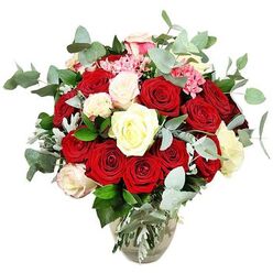 A sumptuous rose arrangement is captured against a crisp white background, showcasing the kind of expert craftsmanship you might find in a studio just off Clerkenwell Green. At the heart of the bouquet lies a generous gathering of deep red and vivid scarlet roses, some tightly furled and others fully opened into lush, layered cups of velvet, creating a sense of warmth and intense devotion. Interspersed throughout are creamy white and soft ivory roses, their petals smooth and luminous, offering a gentle pause between the more fiery tones. A few of these paler roses carry the faintest blush of pink on their edges, like a whisper of affection that softens the overall palette. Tiny clusters of bright pink filler flowers, perhaps mini carnations or berry-like blooms, nestle among the larger roses, introducing a playful spark of colour and a finer, more intricate texture. Encircling and weaving through the flowers, rounded blue-green eucalyptus leaves arc gracefully outward, their matte, silvery sheen and imagined fresh scent adding a modern, soothing counterpoint. Additional deep green foliage, along with some cooler silvery-green leaves, gives the bouquet its full, rounded silhouette and ensures there are no gaps, just an unbroken sweep of petals and greenery. All of this luxuriant growth rises from a clear glass vase with a slightly bulbous base and narrower neck, the stems visible through the water like a tangle of green lines. The arrangement feels ideal for a romantic celebration in Clerkenwell, from an anniversary dinner near St John Street to a surprise delivery to a converted loft, expressing love, admiration, and a sense of occasion.