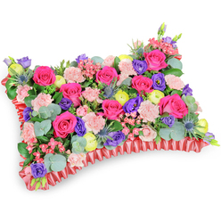 Captured from a slightly raised, three-quarter viewpoint on a clean white surface, this rectangular pillow tribute is a study in careful contrasts and comforting detail-an arrangement we craft at Clerkenwell Flowers for families across Clerkenwell, Farringdon and nearby Leather Lane. The densely packed top is dominated by hot-pink roses, their velvet petals folded in both tight buds and gently opening cups that reflect the soft studio light. Pale pink carnations ripple through the mass, their frilled edges softening transitions between blooms and creating that familiar, tactile ruffled texture beloved in memorial pieces. Scattered energetic notes of lemon-yellow ranunculus brighten the composition like remembered laughter, while clusters of deep purple lisianthus add a cool, contemplative depth. Stiff, silver-blue eryngium thistles punctuate the pillow with a spiky, sculptural rhythm that keeps the eye moving; their bristling form contrasts with the roses' plushness. Interspersed eucalyptus leaves-round, silvery-green and slightly fragrant-lend a calming herbal note and frame the flowers against a backdrop of darker greenery. A pleated, deep-red satin ribbon skirts the perimeter, its folded edges catching light and defining the cushion's silhouette with elegance. The overall effect is intimate and respectful: a vibrant, life-affirming sympathy tribute designed to comfort, to recall joyful colour, and to sit gracefully at a service or graveside in Clerkenwell or St John's Square.