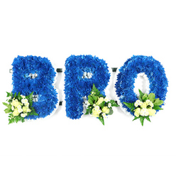 A close, deliberate studio composition shows a three-letter memorial reading BRO, each letter sculpted from dense clusters of royal blue chrysanthemum blooms. Shot at eye level against a pure white backdrop, the letters appear three-dimensional and plush, the tiny chrysanthemum florets layered so the surface reads like a deep blue textile. Strategically placed accent clusters at the lower edge of each character introduce creamy, pale yellow and ivory spray roses - some tightly furled, others easing into bloom - mingled with small white filler flowers such as mini chrysanthemums or baby's breath and a selection of green foliage: broader glossy leaves (salal or lemon leaf) and slender, fern-like fronds that give a soft, feathery counterpoint. A slim white foam frame is visible around each letter and inside the loops, betraying the careful mechanics of a professional funeral tribute.

As a Clerkenwell florist I often recommend this palette for memorials: the deep blue reads as calm and sincere while the ivory roses bring a quiet light. The scent is understated - a green, floral whisper from the roses and foliage rather than perfume - and the even studio lighting highlights the tactile quality of the blooms without drama. This style of tribute, crafted for viewing at a wake, service or graveside, carries a personalised, familial message of respect and remembrance that residents near Exmouth Market, St John's Square or Leather Lane frequently choose when arranging flowers for a lost brother. Clerkenwell Flowers can prepare similar pieces for collection or local delivery in the EC1 area.
