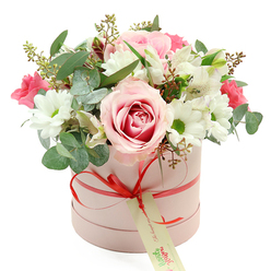 Centred on a clean white backdrop, a chic pastel pink hatbox overflows with fresh flowers, creating a refined yet inviting display that feels right at home in Clerkenwell's creative streets. The cylindrical box, with its smooth, powdery finish, is wrapped near the base with a slim coral-red satin ribbon, knotted into a precise bow at the front. From this bow, a cream rectangular tag with soft green script lettering hangs lightly, adding a personal, almost handwritten touch, as though prepared for a special doorstep delivery. Rising from the box, the bouquet forms a generous domed silhouette, brimming with carefully chosen blooms. A prominent bi-colour pale pink rose, fully opened and luxuriously layered, takes centre stage, its deeper pink heart drawing the eye. Around it cluster white daisy-like chrysanthemums, each flower framed by tidy petals and finished with a bright green centre that adds freshness and clarity. Threaded among them, white alstroemeria show off their elongated, trumpet-shaped petals and fine speckled markings, while smaller deep pink blossoms-similar to spray roses or lisianthus-introduce richer magenta tones that keep the palette from feeling too delicate. The foliage is thoughtfully composed: rounded silver-green eucalyptus leaves curve outward, their soft, dusty surface lending a modern calm, while darker green leaves and feathery sprigs fill any gaps with natural volume. A few tiny green buds and subtle brownish seed heads contribute a gentle, woodland texture. The entire arrangement looks expertly balanced, like something a local florist might prepare for same-day delivery to an apartment near Exmouth Market or a studio off Farringdon Road, ideal for marking a romantic evening, Mother's Day brunch, or a quiet congratulations with understated elegance.