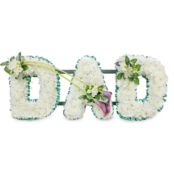 A solemn and lovingly detailed funeral floral tribute spells out DAD in three-dimensional letters, carefully arranged as though ready to be placed beside a coffin or memory table in a Clerkenwell chapel. Each letter is entirely covered with tightly clustered white chrysanthemums, or similar small white button blooms, forming a plush, snowy surface that looks soft to the touch and symbolises purity, respect, and a gentle farewell. Around the perimeter of each letter, a vivid emerald green satin ribbon is pleated into a ruffled border, neatly framing the snowy flowers and offering a fresh contrast reminiscent of the green spaces dotted around Clerkenwell, from Spa Fields to St James's Church garden. Behind the flowers, slim dark bars of the supporting frame peek through, revealing that the tribute is mounted to stand upright as one continuous display. The first D is embellished on its upper left curve with a small, considered cluster of white freesia blooms nestled into rich, glossy foliage, the stems arranged to look naturally grown rather than forced. Two slender, arching green stems extend from this area, sweeping diagonally across towards the central A, where they open out into a striking yet understated spray. Here, two deep purple calla lilies, with their smooth, sculpted trumpets, rest among more white freesia flowers and vibrant green leaves, introducing a ribbon of colour that feels both regal and restrained. The final D carries a complementary spray of freesias and greenery at its upper right, slightly more compact but perfectly balanced. Set against a bright white background, this tribute conveys a moving sense of gratitude, loss, and enduring love for a father, ideal for memorial services and funeral ceremonies in Clerkenwell and the surrounding City streets.