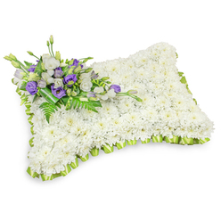 A meticulously formed floral memorial pillow, entirely covered with a dense mosaic of small, pure white chrysanthemum heads, rests on a pristine white surface where a subtle drop shadow gives the cushion a gently raised, three-dimensional presence. The chrysanthemums create a calming, snow-white texture whose close-packed petals read as softness and repose; in person they carry a faint, clean fragrance that evokes chapel lilies and quiet rooms. Encircling the rectangular pillow is a pleated, light green ribbon border-slightly lime in tone-worked into neat overlapping folds that frame and freshen the composition. Diagonally placed at the top-left is a small, artful cluster of accents: white lisianthus with soft, ruffled petals paired with rich purple lisianthus blooms and a few tightly furled buds, the purple providing a measured note of dignity and remembrance. Slender fern fronds and bold dark-green leaves are interwoven through the cluster, while a broad leaf-wrap secures the stems into a composed, respectful unit. The overall presentation is luminous and restrained, photographed to highlight texture and form rather than excess decoration. As a Clerkenwell Flowers-style tribute suited to memorials held at local chapels near Exmouth Market, St John Street or Spa Fields, this pillow is intended to comfort mourners, offering a gentle visual language of peace and enduring care for those arranging funerals or laying flowers at a graveside.