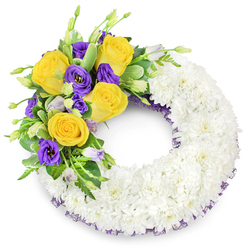 Presented against a clean white field, this circular sympathy wreath reads as both an object of comfort and a small work of floral craftsmanship. The greater part of the ring is composed of tightly clustered, pure white spray chrysanthemums whose dense, layered petals form a smooth, cloud-like surface; the repetition of tiny blooms creates a tactile softness that invites gentle touch and conveys purity. At the upper left quadrant a purposeful cluster of colour rises like a thoughtful exclamation: bright yellow roses in full bloom, their petals spiralled into open cups that suggest warmth and friendship, paired with luxurious royal purple lisianthus (Eustoma) whose ruffled edges and satiny sheen introduce a cool, contemplative counterpoint. A scattering of unopened lisianthus buds in pale lavender and green adds texture and the promise of ongoing life. Supporting greenery - glossy rounded leaves, delicate fern fronds, and light-variegated foliage - frames the accent and provides depth, separating the vivid cluster from the monochrome base. Along the inner and outer edges, a narrow trim of deep purple mesh or crinoline peeks out from beneath the chrysanthemums, a subtle detail that ties the wreath's palette together. The wreath's central opening preserves the traditional circular form, while the balanced asymmetry of the accent makes the design feel intentional and tender. In soft daylight the arrangement appears serene: the white chrysanthemums quiet the eye, the yellow roses offer warmth, and the purple lisianthus sustain dignity. Designed for memorial or bereavement settings, this wreath expresses calm remembrance with thoughtful floristry and understated beauty, suitable for local services in and around Clerkenwell and neighbouring Farringdon.