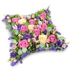 This image shows an elegant floral pillow tribute, created in a soft rectangular cushion shape that conjures thoughts of peaceful rest and gentle support. The entire surface is densely covered with fresh blooms, with no gaps, giving the arrangement a plush, almost fabric-like appearance. Dominating the design are classic roses in shades of vivid fuchsia, gentle powder pink, and creamy lemon-cream, many open to reveal their swirling, velvety petals. Nestled among them are deep royal purple lisianthus flowers, their ruffled forms adding depth and a mellow, contemplative tone. Tiny lavender and lilac filler flowers, similar to statice or limonium, thread between the larger heads like fine embroidery, while petite blush pink spray roses soften any strong lines. Around and beneath the blossoms, cool silvery eucalyptus leaves mingle with darker, ferny greenery, building a rich, layered green base that frames the colours beautifully. Set against a pure white background with a subtle shadow beneath, the tribute appears freshly prepared and ready to be delivered to a local Clerkenwell chapel, a memorial gathering near Farringdon, or a quiet family farewell just off Clerkenwell Road. The composition feels calm and reassuring, a thoughtful funeral or sympathy cushion designed by a florist who understands how flowers can carry love, gratitude, and remembrance when families in EC1 seek a gentle way to say goodbye.