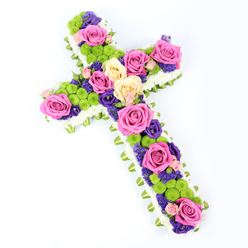 Set against a spotless white backdrop, this cross-shaped floral tribute unfolds like a tapestry of colour and texture, designed with the same quiet care you might feel walking past the churches and historic streets of Clerkenwell. The cross is first defined by a thick, pillowy border of tightly packed white spray chrysanthemums, each tiny floret pressed close to its neighbour, creating a soft, almost feathered edge. Just behind this snowy halo, a neatly folded light green ribbon or collar of foliage encircles the entire shape, slipping into view here and there to add a refined, tailored finish and a gentle suggestion of new growth. Within this frame, the heart of the cross is an intricate composition of fresh blooms. Large roses in hot pink and creamy pale yellow are arranged in clusters along the vertical and horizontal beams, their full, layered petals opening generously as if in a final, fragrant blessing. Nestled among them are bright lime-green button chrysanthemums, their rounded, textured heads giving the design a lively, almost playful rhythm. Small deep purple and violet flowers, likely lisianthus or statice, wander between the larger blossoms, deepening the palette and creating pools of shadowy colour that draw the eye inward. The cross is photographed at a slight diagonal from top left to bottom right, enhancing its three-dimensional form and giving it a gentle sense of movement, as though it has just been placed for a service. The lighting is bright but soft, revealing each detail without harshness. This is a thoughtful Christian funeral arrangement, ideal for a memorial in Clerkenwell or nearby Farringdon, expressing faith, remembrance, and a tender hope through its harmonious blend of pink, green, and purple flowers.