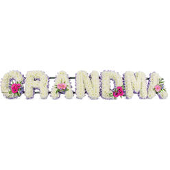 In this tender funeral flower tribute, the word GRANDMA is shaped from broad, plush letters, each one fully carpeted with compact white chrysanthemums that create a smooth, almost cloud-like surface. The blooms are tightly clustered, giving the impression of gentle strength and reliability, much like a grandmother's embrace remembered on quiet walks through Clerkenwell's side streets. A continuous lilac ribbon outlines every letter, its cool, satin sheen catching the light so that the edges seem to glow softly, adding refinement without overwhelming the calm white base. At three thoughtful points along the word, the white chrysanthemums part to reveal small, rounded sprays of mixed flowers. Pink gerberas with their open faces and tiny central discs sit alongside delicate pink roses, some in bud and some just unfurling, tucked amongst glossy green foliage that introduces movement and depth. These little islands of colour feel like treasured memories - bright moments within a life honoured with respect. The lighting is even and gentle, allowing each petal, leaf, and ribbon fold to be clearly defined, as if the tribute were resting in a peaceful Clerkenwell chapel or a quiet corner near Exmouth Market before a service. Though completely still, the design seems to breathe with emotion: a loving statement created with care by skilled hands, ideal for families in Clerkenwell wishing to say goodbye with grace. It is a floral arrangement that balances purity, softness, and personality in a single, meaningful word.