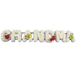 The image shows a lovingly crafted GRANDMA funeral tribute laid out in a straight horizontal line, photographed against a stark white backdrop so that the detail of every flower can be appreciated. Each bold capital letter is entirely filled with tightly clustered white chrysanthemums, their small, domed petals overlapping to form a thick carpet of blooms that looks soft, cool, and almost velvety to the touch. Just peeking out beneath the white, a fine trim of pale lavender flowers skims along the lower edges of each letter, like a shadow of colour outlining the shape and giving the design a gentle lift. On the left, the G carries a cluster of three deep red roses at its lower curve, the petals spiralling in rich layers and surrounded by tidy dark green foliage. The first A is decorated with two striking yellow-green orchids resting gracefully on its crossbar, their patterned throats tinged with reddish tones, suggesting individuality and cherished stories. The R, N, and D remain pure in white and lavender, steady and composed. Near the right side of the arrangement, the M holds another trio of red roses cupped within its lower right dip, echoing the first cluster and balancing the composition, while the final A repeats the pair of vibrant green orchids. A slim, dark metal rail links the letters discreetly along the top, giving enough support for display at a church or crematorium in and around Clerkenwell, from Farringdon Road to Exmouth Market. The tribute feels both traditional and contemporary, offering a serene, floral expression of love and remembrance for a grandmother who meant the world.