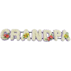 Set against a clean white backdrop, a lovingly crafted funeral tribute spells GRANDPA in large, three-dimensional block letters made entirely from white chrysanthemums. Each letter is densely packed with small, daisy-like blooms, their soft petals overlapping to create a plush, cushiony texture that looks gentle to the touch and evokes calm, respectful remembrance. Along the base of each letter, a subtle band of pale lilac or lavender ribbon peeks through, adding a cool wash of colour that outlines the word without overwhelming the simplicity of the white flowers. Discreet dark green bars connect and support the letters, keeping the structure secure for display at a crematorium chapel or church service in Clerkenwell, perhaps near Farringdon or Hatton Garden. Carefully placed floral sprays provide moments of vivid colour and emotion: on the G and D, clusters of deep red anthuriums with their distinctive, glossy, heart-shaped spathes and pale central spadices nestle into rich green leaves, symbolising enduring love and the warmth of family bonds. The first A and the last A are softened with sprays of yellow-green orchids, their slender stems and intricate blooms giving a feeling of refinement and grace. Additional foliage curls naturally around these feature flowers, adding depth and gentle movement, as if the letters are breathing with life and memory. The result is a personalised GRANDPA floral name tribute that conveys tenderness and gratitude, offering families in Clerkenwell a serene, beautifully detailed way to say a final thank-you to a cherished grandfather.