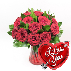 A rich, romantic arrangement of deep red roses appears to float in front of a pristine white backdrop, the kind of passionate display a Clerkenwell florist might prepare for a candlelit proposal near St John's Square. The bouquet features roughly a dozen or more roses in a sumptuous crimson shade, their petals thick and velvety, some blooms cupped and opening, others fully spread in generous rosettes. Nestled among them, fresh green foliage adds texture and depth, the leaves broad, glossy, and layered so that the reds and greens weave together like fabric. All of this is gathered into a ruby-red glass vase, clear yet tinged with colour, its sides subtly fluted so that vertical ridges catch the light in slender highlights. The vase's rich tone echoes the roses, giving the arrangement a cohesive, almost jewel-box quality. In the lower right foreground, leaning toward the flowers and partly overlapping them, a shiny red heart-shaped foil balloon steals a little of the spotlight. Its surface gleams with bright reflections, and at its centre, the words "I Love You" curve in elegant white cursive script. A stylised silver arrow, reminiscent of Cupid, appears to pierce the heart on a gentle diagonal, adding a playful, storybook touch to the composition. The plain white background throws every colour into sharp focus, making the reds seem even deeper and the foliage more vibrant. Together, vase, roses, and balloon form a classic romantic gift, ideal for same-day delivery around Clerkenwell when someone wants to send a message of devotion that feels both joyful and sincere.