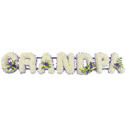 The image shows a carefully composed funeral flower arrangement spelling GRANDPA in bold, upright capital letters that stretch evenly across a white background, as if placed on a table in a quiet Chapel Street room in Clerkenwell. Each letter is built on a hidden frame and completely covered with compact white chrysanthemums or carnations, so tightly massed that no gaps are visible. The flowers' rounded petals create a plush, cushioned texture, like a soft white quilt of remembrance laid gently across the word. Within this uninterrupted field of white, the florist has added considered accents of purple and lavender flowers, accompanied by sprigs of glossy green foliage. These coloured touches appear in key points of the design - cradled at the base of the G, brightening the centre spaces of both As, and softening the upright and loop of the P - bringing gentle depth rather than distraction. A slim, dark green bar runs underneath all seven letters, largely hidden by leaves and blooms, providing the structure needed to move the tribute from the shop to a funeral car or a service along Clerkenwell Road. The combination of crisp white and tranquil purple gives the piece a serene, almost contemplative feel, suitable for resting beside a coffin, at the foot of an altar, or near a photograph of the grandfather being honoured. Crafted with patience and care typical of Clerkenwell Flowers' work, this tribute offers families a personalised way to say goodbye, with each blossom seeming to carry a quiet message of love, respect, and enduring memory.