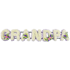 The image shows a beautifully composed funeral flower tribute spelling out GRANDPA in bold, three-dimensional letters, created with the kind of care a local Clerkenwell florist would bring to a family ceremony at a nearby chapel or cemetery. Each letter is formed from a thick blanket of small white chrysanthemums or similar dainty blooms, packed closely together to create a velvety, cushion-like surface that looks soft enough to rest a hand upon. Around the edges, a neat outline of fine purple and lilac flowers traces each curve and corner, offering a cool, lavender-toned frame that gently separates the letters from the clean white backdrop. Throughout the tribute, thoughtfully placed accent sprays add colour and personality: on the 'G' and the first 'A', delicate white lily-style flowers stretch gracefully above clusters of pink and fuchsia blossoms, their tones deepening into amethyst and plum purples. The 'D' and another 'A' mirror these groupings, some of them brightened with small lime-green button chrysanthemums and fresh foliage, like tiny garden scenes reminiscent of hidden courtyards off Clerkenwell Green. A narrow, dark green bar runs along the back, almost invisible yet quietly holding the letters in perfect alignment, ready to sit along the front of a coffin or at the head of a graveside tribute. The lighting is soft and even, without harsh shadows, allowing the gentle sheen on the petals and the contrast between white, purple, and pink to stand out. Overall the piece speaks of respect, gratitude, and a lifetime of memories, a floral embrace for a cherished grandparent.