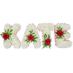 This personalised floral name tribute spells KATE in large, sculpted block letters, each one formed from a dense, cloud-like mass of immaculate white chrysanthemums that give the letters a soft, tactile presence. Around the outer edges a pale pink satin ribbon - pleated and neatly finished - traces each contour, introducing a gentle blush that complements the white blooms. On every letter a single scarlet rose provides a dramatic focal point: the K bears its rose on the lower portion of the left vertical stroke, the A's rose sits at the apex of its triangular peak, the T's rose anchors the left base of the horizontal crossbar, and the E's rose is placed on the middle horizontal arm, each rose cradled by clusters of lush green foliage including glossy broad leaves and fine ferny sprigs. The overall effect is quietly elegant and respectfully expressive, suitable for a funeral, memorial service, or a commemorative display. Soft, even lighting accentuates the chrysanthemum's texture and the roses' velvety depth while the arrangement's restrained scent of roses lends a gentle sense of presence. Made with care in our Clerkenwell studio near Exmouth Market, this bespoke tribute reflects a local florist's attention to proportion and meaning; Clerkenwell Flowers offers thoughtful handling and can prepare similar personalised tributes for services at nearby venues such as St John's Church or the crematoria serving Farringdon and the surrounding area.