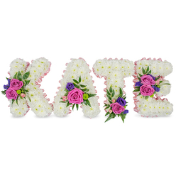 This lovingly composed name tribute spells KATE in four freestanding letters, each letter built from a thick, luxurious carpet of snowy white chrysanthemums that create a plush, seamless surface. A soft pink satin ribbon outlines every contour and inner space, giving the letters a faint halo of blush against the studio-bright white background. Vibrant accent groupings - dominated by deep fuchsia-pink roses, small pastel pink spray roses, and touches of deep purple or blue blooms such as statice or petite lisianthus - are arranged with a florist's careful eye to provide instant contrast and visual warmth. Lush green elements, from feathery fern fronds to smooth eucalyptus leaves, tuck around the blooms to add depth and a natural, calming palette. Placement is intentional: a cluster on the diagonal of the K, one at the lower right curve of the A, an anchoring bunch at the base of the T, and two thoughtful groupings on the middle and lower bars of the E, which together create a gentle flow across the whole name. The texture reads soft and tactile, the roses velvety against the chrysanthemum cushion, the scent subtle and consoling. Appropriate for a funeral service, memorial, or celebration of life, this personalised floral arrangement conveys sympathy, remembrance, and love. As Clerkenwell-based florists we craft these pieces with local families - often delivering to chapels near Exmouth Market, services around Smithfield, or private remembrances in St John's Square - ensuring the tribute sits respectfully within the service.