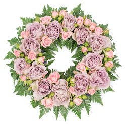 The image presents a carefully crafted memorial wreath photographed from directly above, resting against a pure white background so that its tranquil colours and textures stand out clearly. Forming the ring is a dense circle of large, fully opened roses in dusty lavender and antique mauve, their petals unfolding in soft spirals that suggest both fragility and strength. Threaded between them are smaller blush pink spray roses and tiny rosebuds at different stages of opening, adding gentle movement to the design, as if the flowers were caught between one breath and the next. Beneath and around the blooms lies a solid base of dark emerald fern foliage, the fronds overlapping in feathery layers that frame the pastel roses with a calm, grounding green. Slight variations in tone, with deeper buds and shadowed leaves tucked between the flowers, lend depth and a natural, garden-like feel, reminiscent of the pocket parks and planted squares around Clerkenwell and Farringdon. The circular shape is even and harmonious, the inner and outer edges of the wreath cleanly defined, creating a sense of order and quiet dignity so often sought for funeral tributes and celebration of life services. Light falls evenly across the arrangement, highlighting the velvety texture of each petal and the fine patterning of the fern leaves. The overall impression is one of serene beauty and compassionate thoughtfulness: a wreath that might be delivered to a Clerkenwell chapel or family home as a floral embrace, honouring memories, offering comfort, and expressing sympathy when words feel insufficient.
