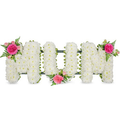 Centred on a clean white backdrop, a lovingly crafted floral tribute spells out the word MUM in broad capital letters, each one structured on a slim dark green frame that peeks out only at the base. The letters are completely carpeted with small white chrysanthemum blossoms, placed tightly together so they resemble a plush, snowy blanket. Their petals layer softly over one another, giving the arrangement a velvety, almost quilted texture that feels calm and soothing. Delicate accents of colour are woven in with intention rather than excess. The first M carries a flourish of pink at its upper left corner: a striking fuchsia rose in full bloom, surrounded by smaller pale pink spray roses, tiny buds, and arching fern fronds that add movement and a fresh, woodland green. The central U is gently cradled by a similar cluster at its lower centre, the pink and fuchsia tones drawing the eye to the heart of the word. On the final M, a mirror image sits at the upper right, completing a graceful diagonal of colour from left to right. Light falls evenly across the flowers, picking up subtle shadows in the chrysanthemum centres and the glossy finish of the leaves, so every texture is visible. The overall effect is one of tender devotion-a classic MUM tribute of the kind often chosen by Clerkenwell families for funerals at local churches or crematoria, or as a graveside token after a farewell. It quietly expresses love, gratitude, and remembrance without overpowering the setting.